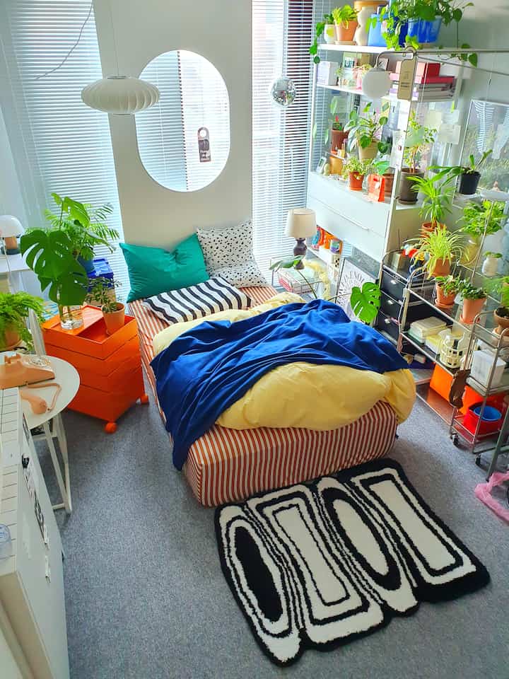 白とグレーの調和が美しいワンルームの寝室、青いベッドカバーと多様な植物、パターンラグが配置された雰囲気のある空間