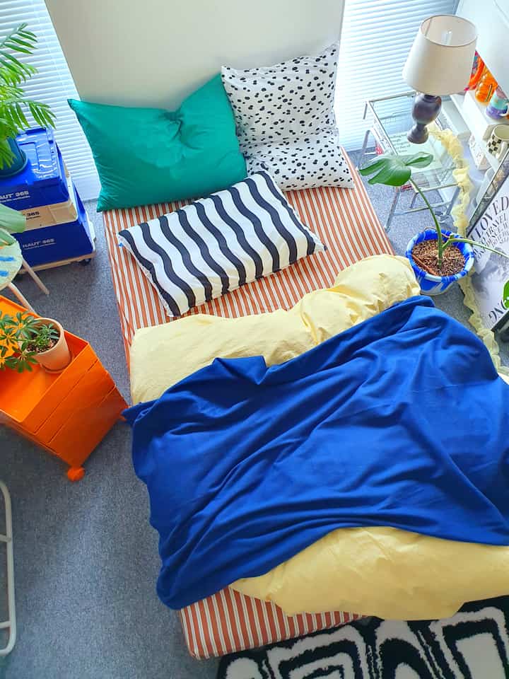 Natural-toned studio bedroom featuring an orange storage unit and bed with patterned pillows and a blue blanket in a cozy, plant-enhanced setting.