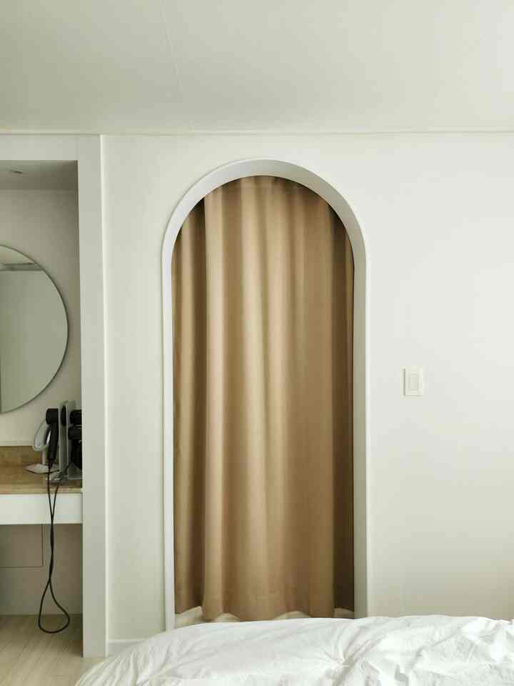 White and beige toned bedroom featuring an arched doorway with beige blackout curtain and a vanity, creating a simple space