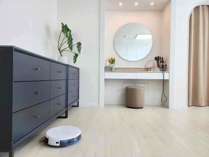 A minimalistic small bedroom in white and dark brown tones, featuring a large dresser on the left and a vanity with a round mirror and stool centrally placed