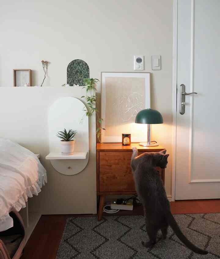 Natural modern bedroom with white walls and bed, wood-tone nightstand, green table lamp, and a cat exploring