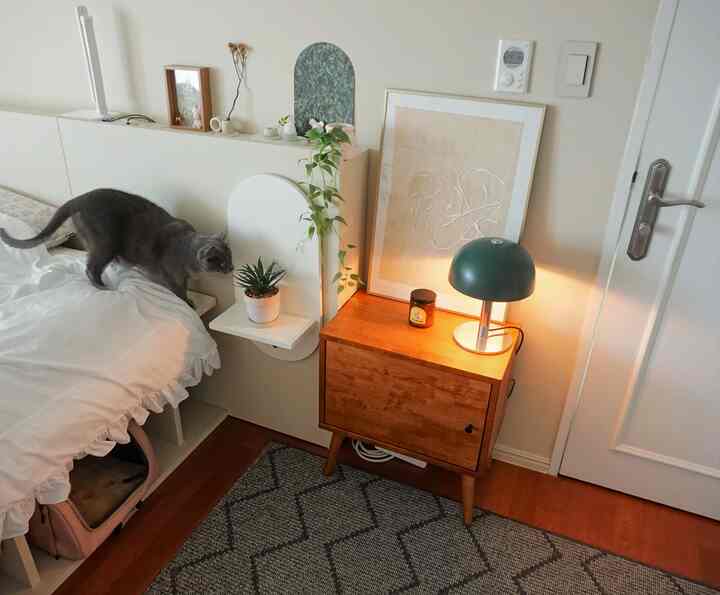 A cozy bedroom in white and brown tones featuring wooden nightstand with green mood lamp and plants, and a gray cat on the bed
