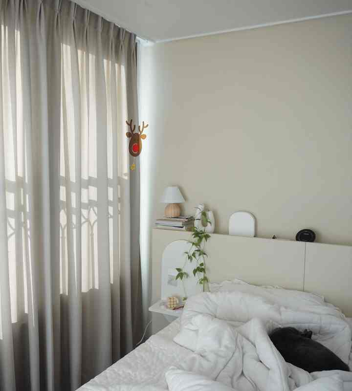 Cream-toned bedroom with blackout curtains by the window and a cat resting on the cozy bed, conveying a soft and calm atmosphere