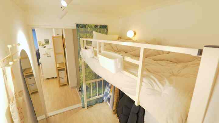 A small loft bedroom with bright ivory and beige tones, featuring wall-mounted light, curtain, and organized storage under the bed in a minimal style