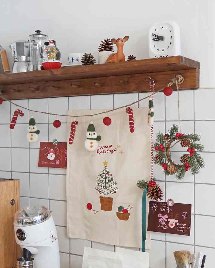 White tiled kitchen space with brown wooden shelf featuring Christmas garland and posters, creating a warm festive atmosphere