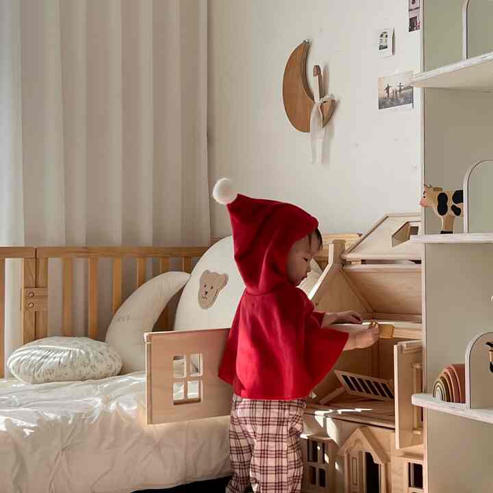 アイボリーのカーテンと木目調の家具が配置された子供部屋、赤ちゃんがドールハウスで遊ぶ温かみのある空間