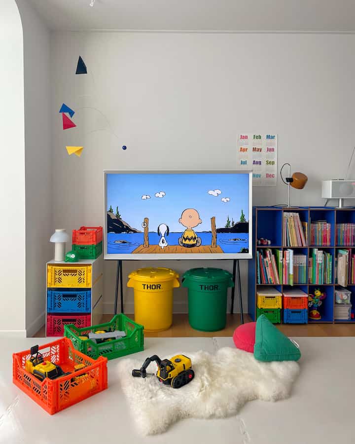 Bright white-walled living room with wood flooring, colorful storage bins, toys, and a TV creating a child-friendly space