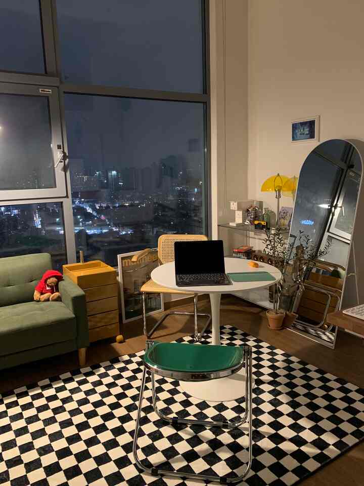 Mid-century modern studio apartment featuring a green sofa, white round table, large floor mirror, and yellow lamp with night city view
