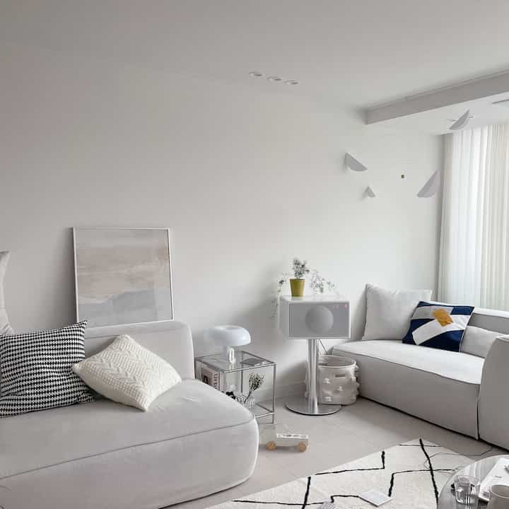 Bright white-toned living room featuring two sofas, a picture frame, table lamp, cushions, and rug in a modern interior