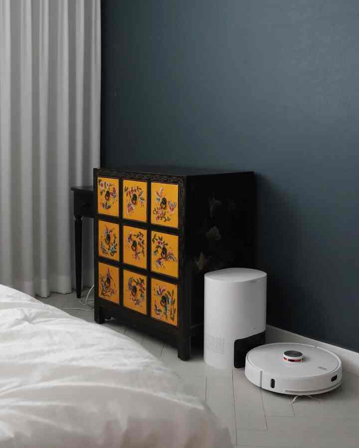 ホワイトの寝具とカーテンがあるブルートーンの寝室、伝統模様のキャビネットとロボット掃除機が配置されたモダンな空間