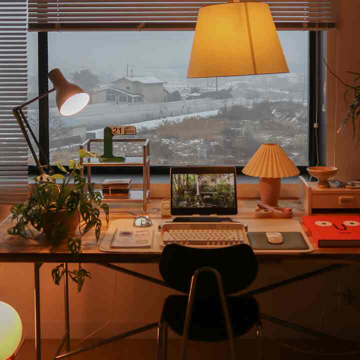Warm yellow lighting illuminates a natural wood desk with assorted items in a home office, large window showing snowy winter landscape outside