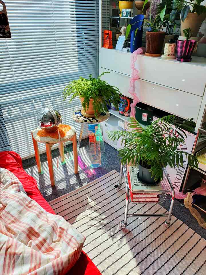 Bright sunlit bedroom with red bedding, wood-tone stools, and lush plants arranged around the space