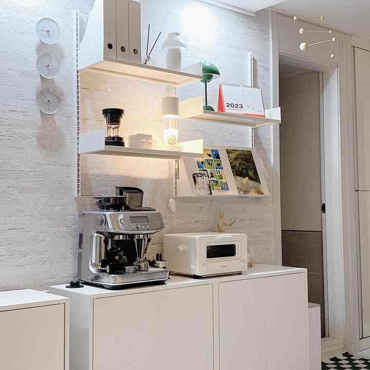 White-toned living room wall featuring floating shelves, cabinets with a coffee machine and toaster, creating a minimal and neat home cafe space