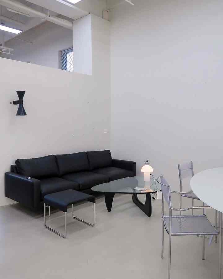 Modern living room with white walls and a dominant black leather sofa, featuring a glass coffee table and a stool