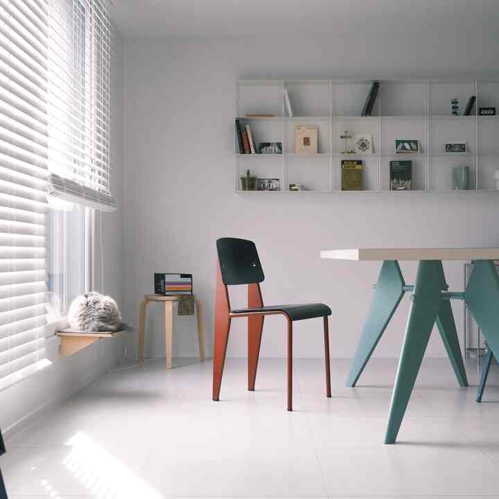 White and wood tone living room with blinds-covered window featuring a cat on a wooden shelf and Vitra dining table and chair in a clean setting