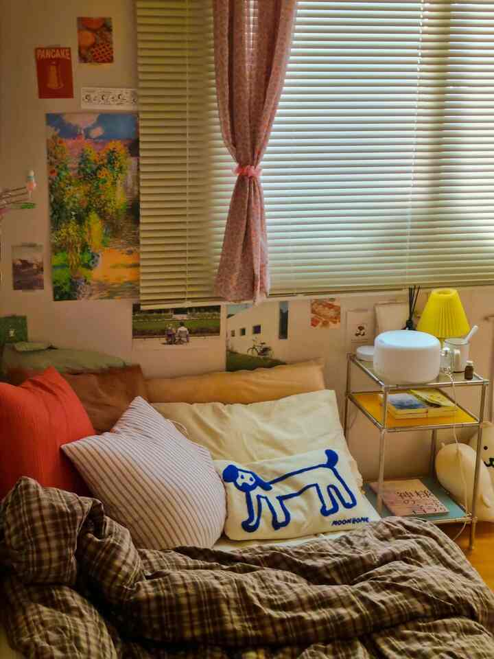 Natural-toned small bedroom with pink curtains and blinds on window; cozy space featuring bed pillows and a side table with lamp
