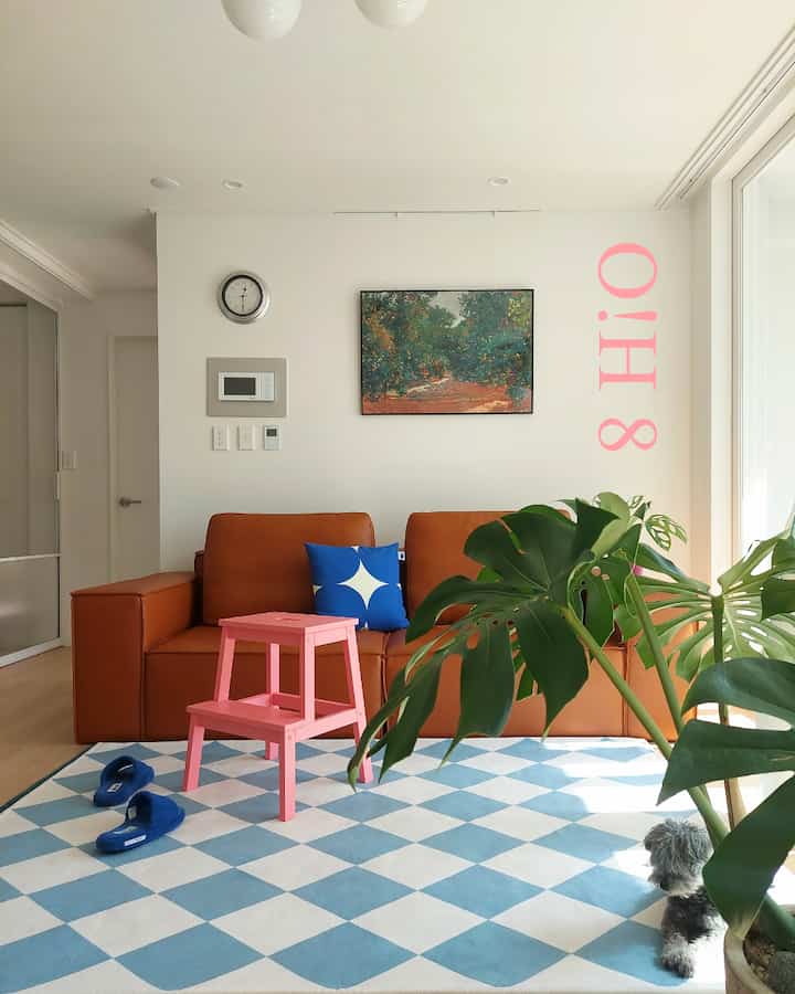 Beige modular sofa with blue cushion and pink stool in living room, cozy space with large plant and small dog