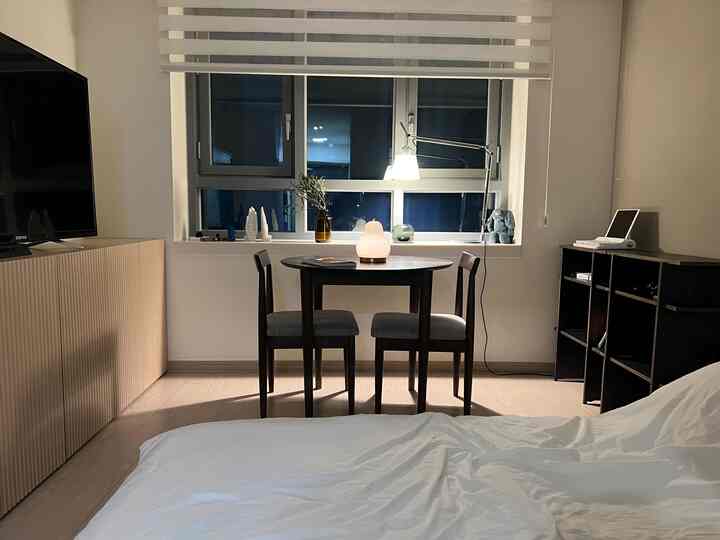 A white and natural wood tone studio apartment featuring a round dining table and single bed, creating a cozy and clean atmosphere