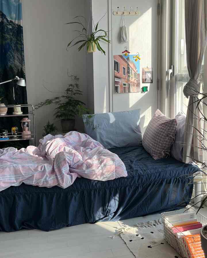 明るい白い壁と濃い青の寝具が調和したワンルームの寝室、植物とポスターが配置されて自然で落ち着いた雰囲気のインテリア