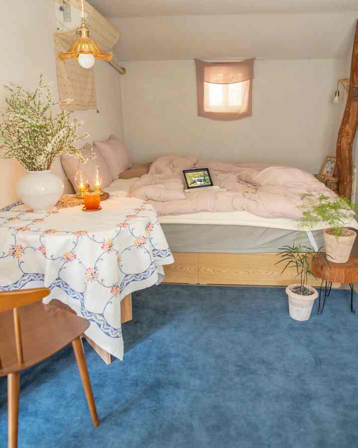 ブルーのカーペットとベージュの寝具がある縦長の寝室、木製の椅子と植物が配置されたナチュラルな雰囲気のインテリア