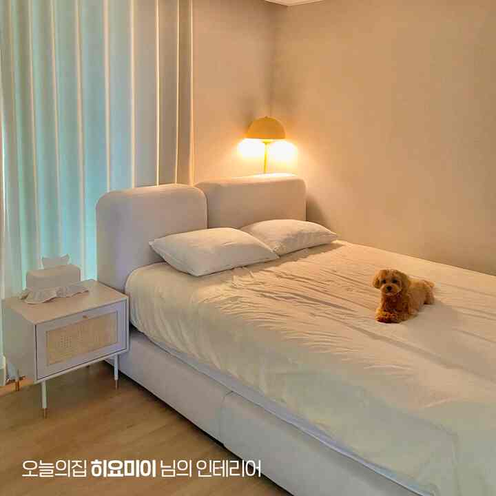 White and natural tone bedroom featuring a bed and nightstand centrally placed, with a yellow floor lamp and a small dog adding cozy atmosphere
