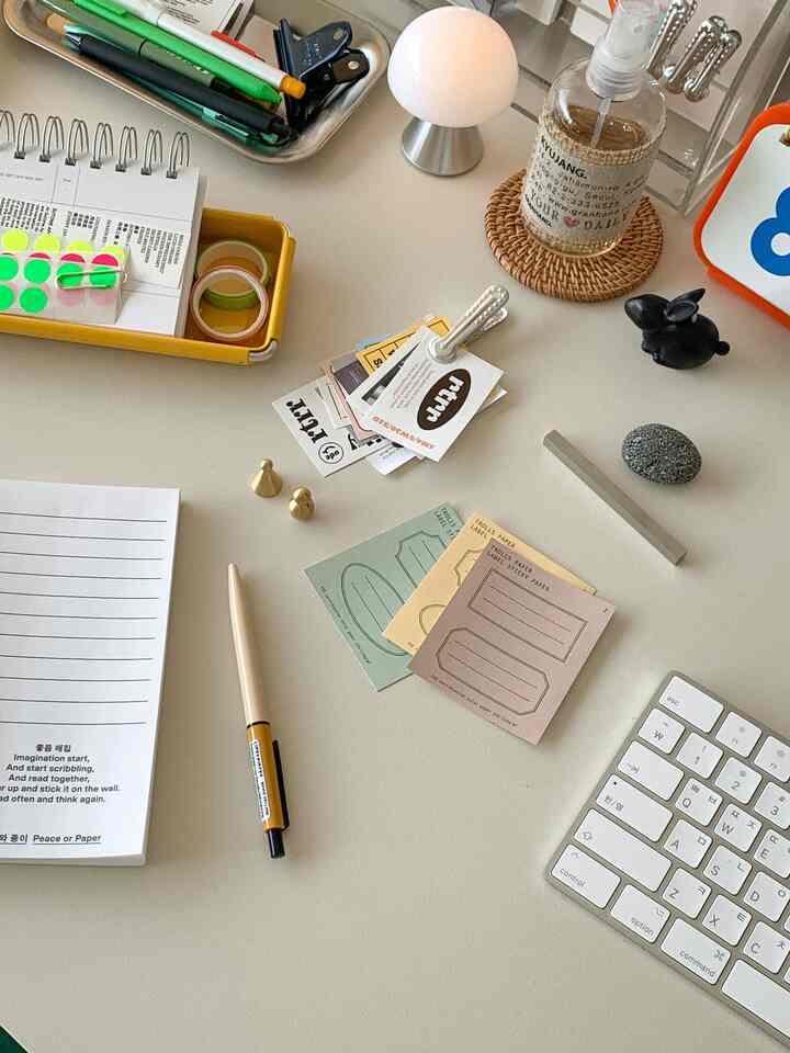 White-toned desk featuring keyboard, pens, stickers, mood lamp, and stationery items arranged neatly in a home office space