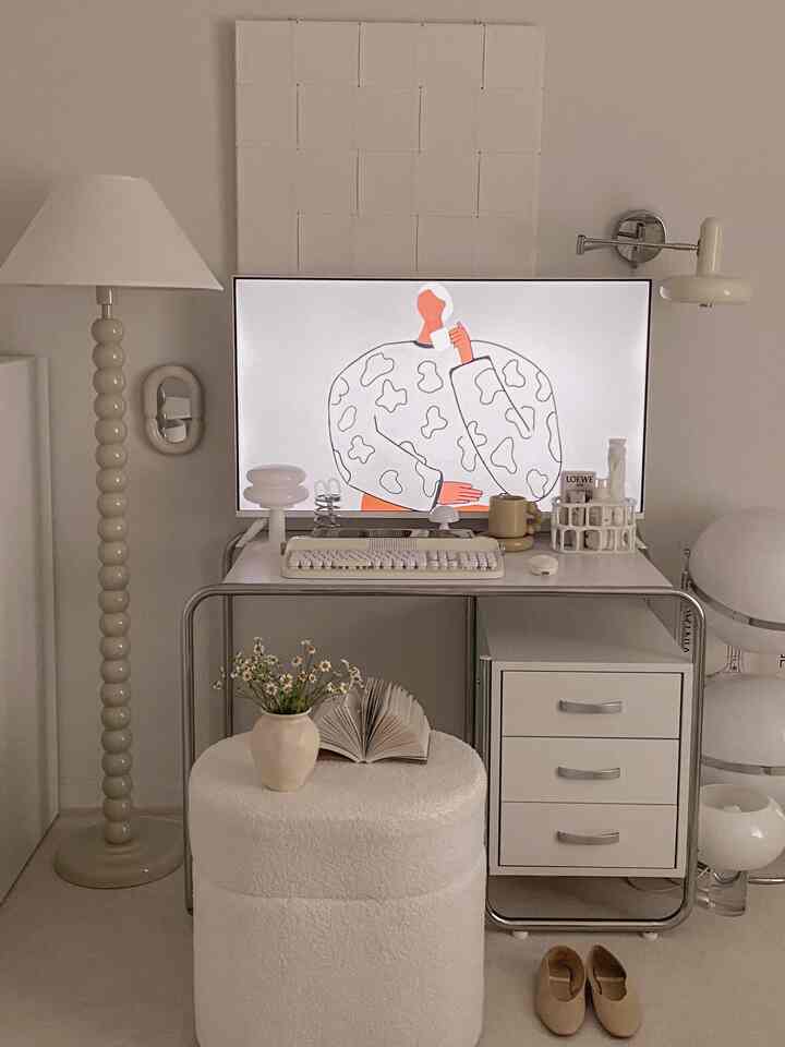 Modern home office in white and ivory tones featuring a TV stand desk, table lamps, and a cozy stool with a book and vase