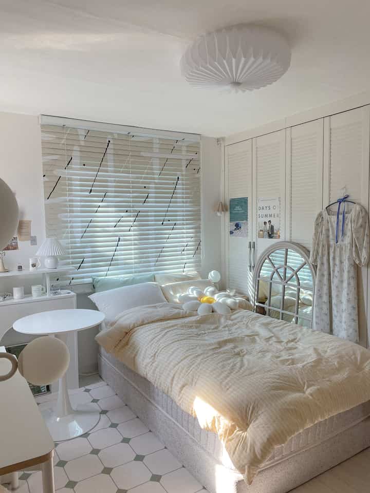 White and beige toned bedroom featuring art-patterned blinds, a large bed, round table, and wardrobe with a warm, simple atmosphere