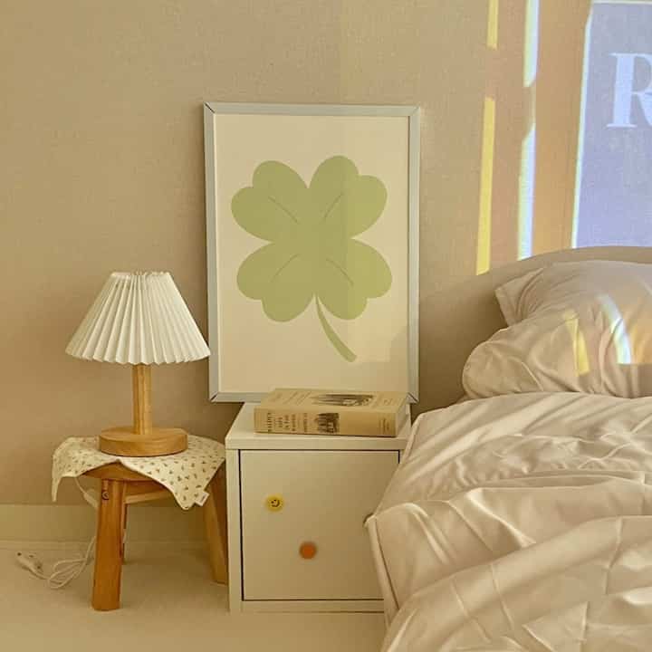Beige and white toned bedroom featuring wood-tone stool and table lamp, with a four-leaf clover poster creating a cozy atmosphere