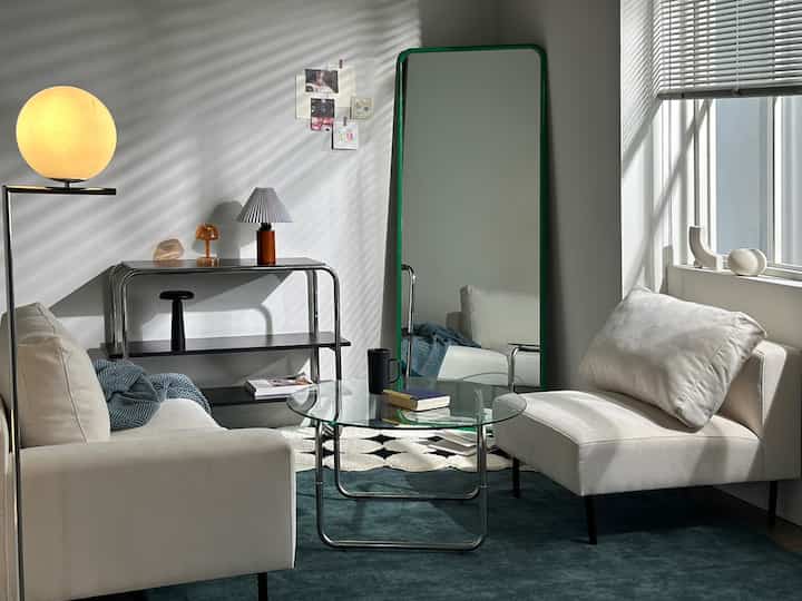 White-walled studio living room with Mid-Century Modern furniture, featuring beige sofa and green-framed full-length mirror creating a cozy atmosphere