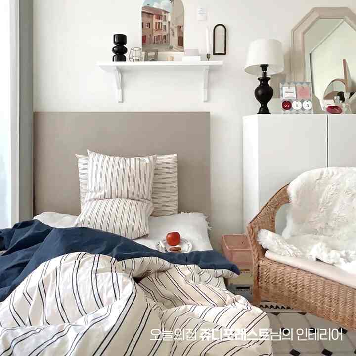 白とベージュの色調の寝室、ストライプの寝具と枕が置かれたベッドと籐のアームチェアが配されたシンプルでナチュラルなインテリア