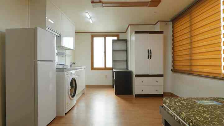 Compact studio apartment with brown wood floor and white walls featuring kitchen appliances and a single bed