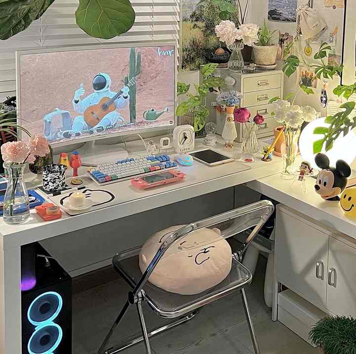 White and gray toned compact home office featuring desk, monitor, character cushion, various plants, and ambient lighting in cozy workspace