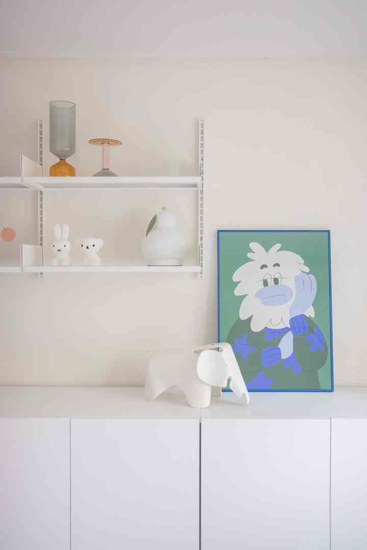 White-toned bedroom or living room space featuring a white cabinet, shelves with cute decor items, and a blue-framed artwork, creating a clean and natural atmosphere