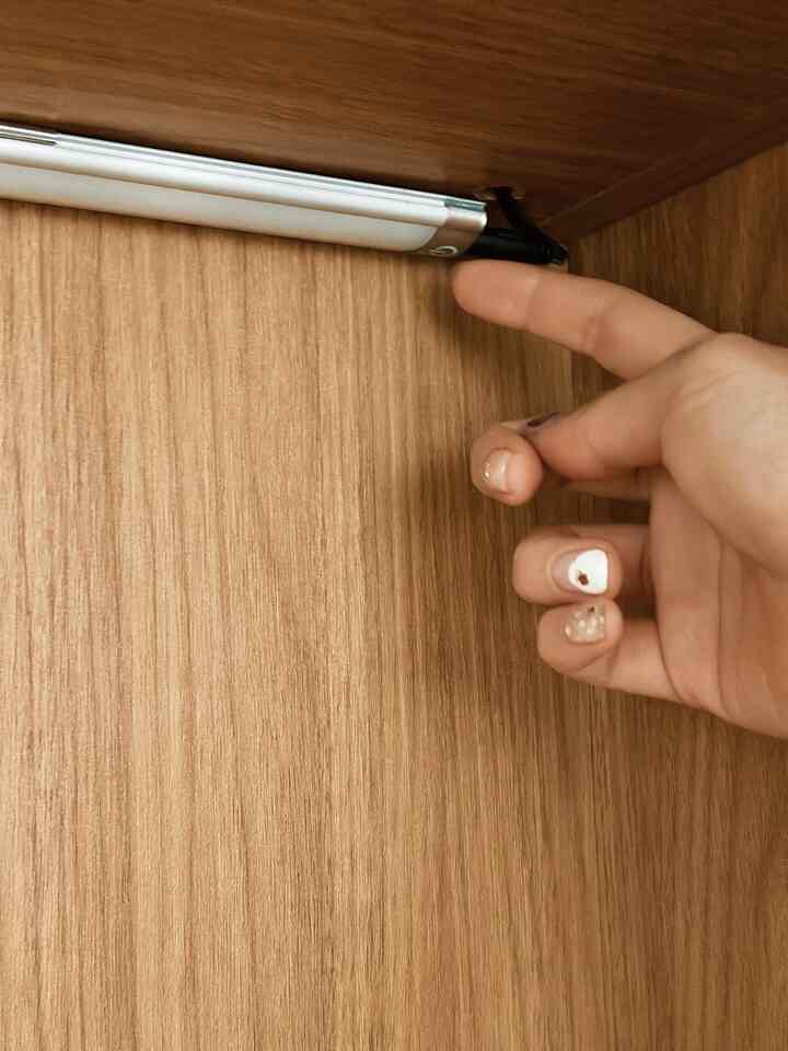 A hand touching a slim touch-activated LED light installed inside a wood-toned shelf