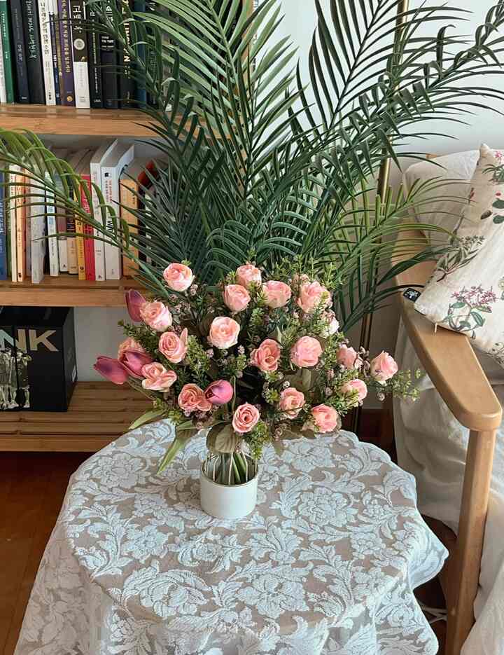 Natural-toned living room featuring wooden armchair with floral cushion and pink artificial flower bouquet on white lace tablecloth-covered round table