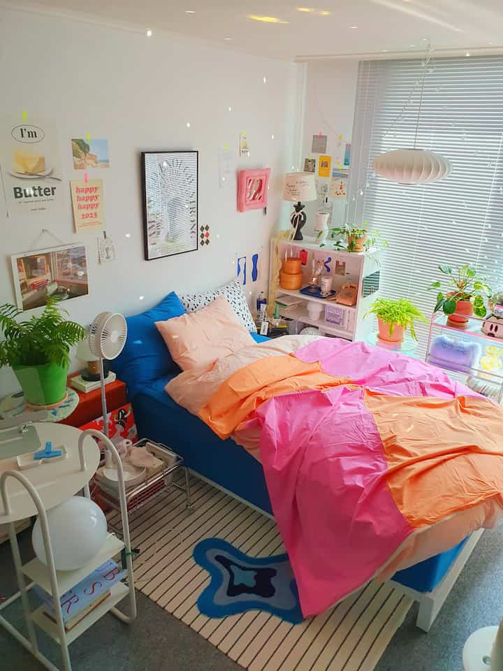 A natural and compact studio bedroom with white walls and blinds, featuring a blue bed with bright pink and orange bedding accents