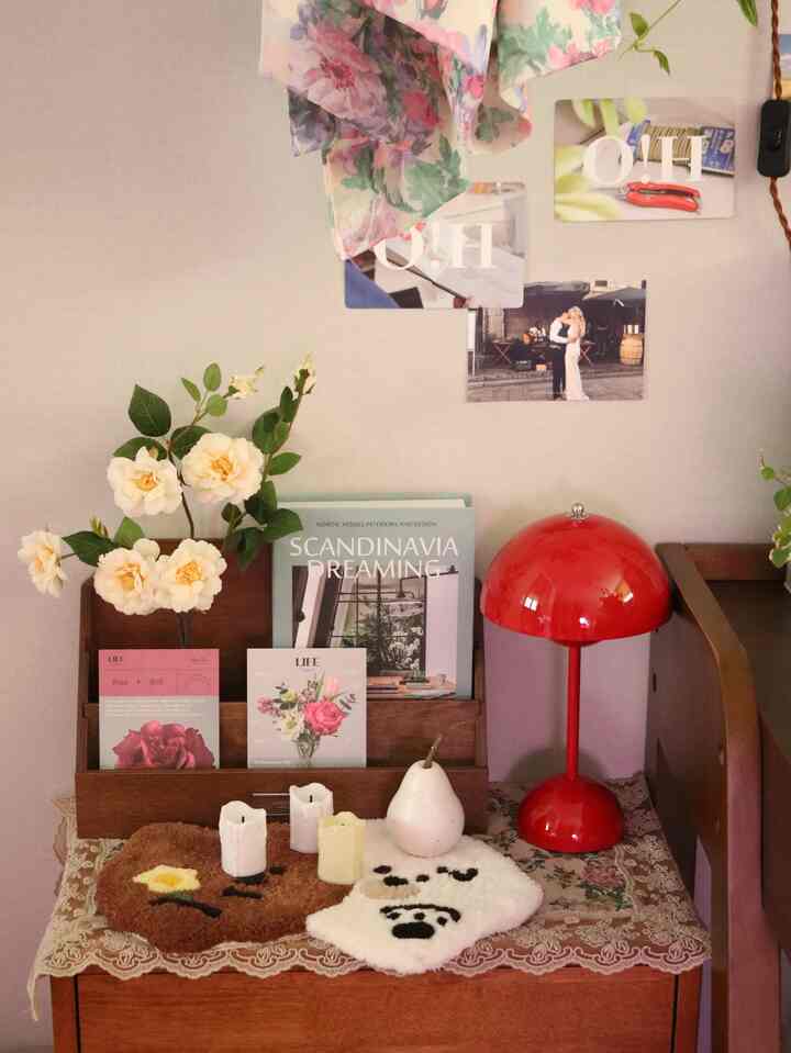 Cozy space with light beige walls featuring a wooden nightstand topped with a red table lamp, vase with flowers, paper storage box, and decorative objects