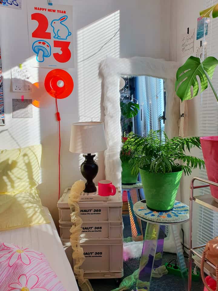 Bright bedroom space in white and green tones featuring plants, a furry-framed mirror, and a table lamp with a cheerful atmosphere