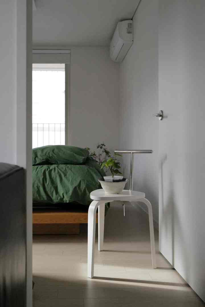 White and green toned bedroom featuring wooden bed with green duvet cover and a white stool with plant, creating a simple natural space