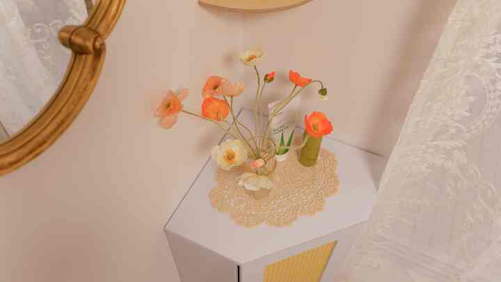 ベージュ色の壁のコーナーにラタンとゴールド素材のコーナーキャビネットが置かれ、ゴールドの額縁鏡と花の装飾がある雰囲気のあるインテリア