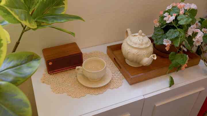 Cozy home cafe balcony with cream walls and white cabinet, featuring wooden tray, teacup set, Bluetooth speaker, and pink flowering plant