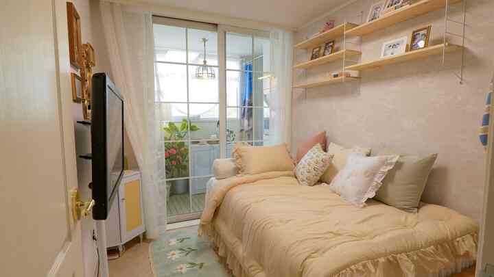 ベージュと白の色調のワンルーム寝室、ベランダに繋がるソファベッドと壁棚、レースカーテンが配置された雰囲気のあるインテリア