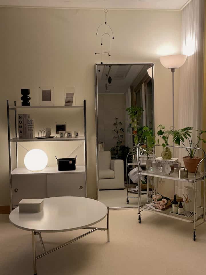 White and silver toned living room featuring a round table, silver frame full-length mirror, mobile shelves with plants, and warm lighting creating a cozy atmosphere