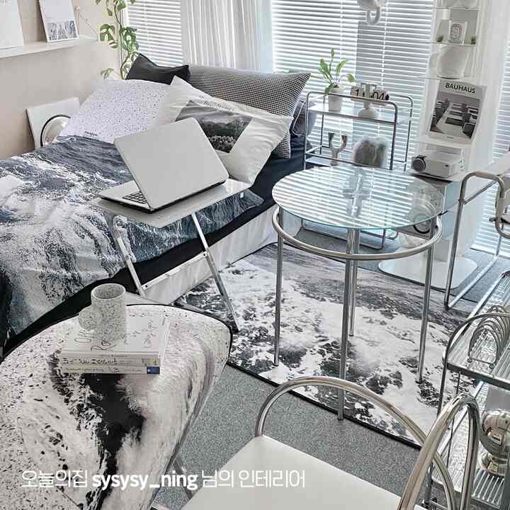 グレーと白の色調のワンルームリビング、ソファベッド、丸いガラステーブル、ブラインド、ラグが調和したモダンでミニマルなインテリア