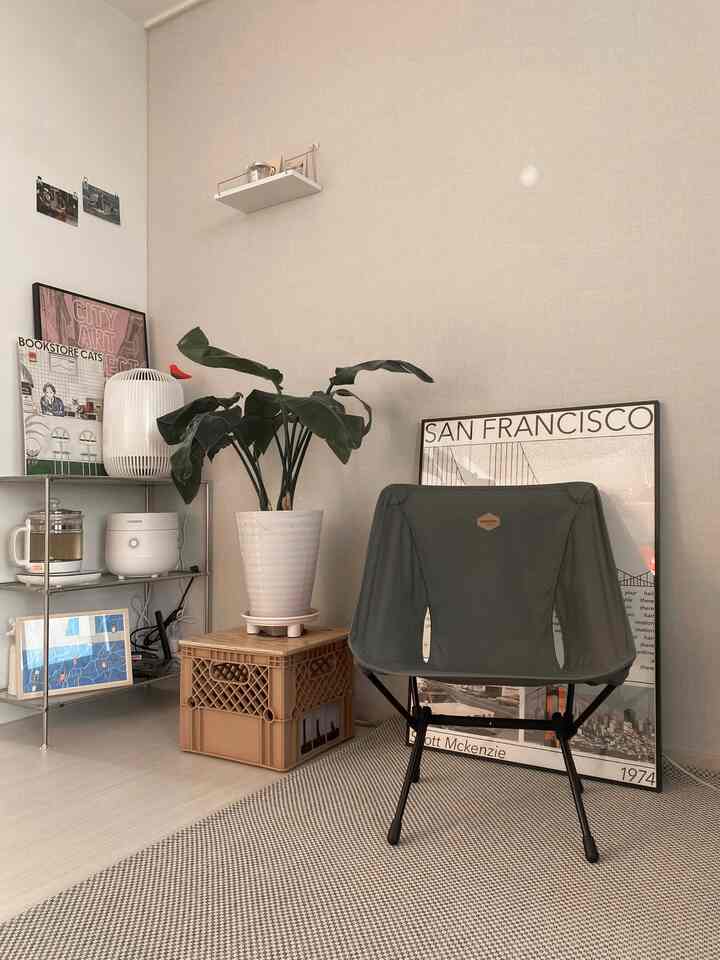 Beige-toned wall and floor with gray camping chair, modular shelf, plant, and posters arranged in cozy living room interior