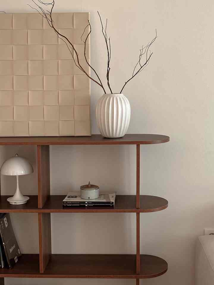 Warm beige and brown toned living room with simple wooden shelf featuring a decorative vase with twigs and table lamp