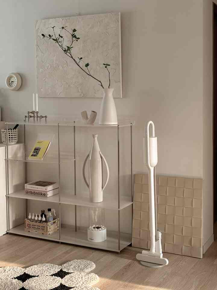 Natural tone living room space featuring transparent shelves and a white cordless vacuum cleaner in a minimal modern interior
