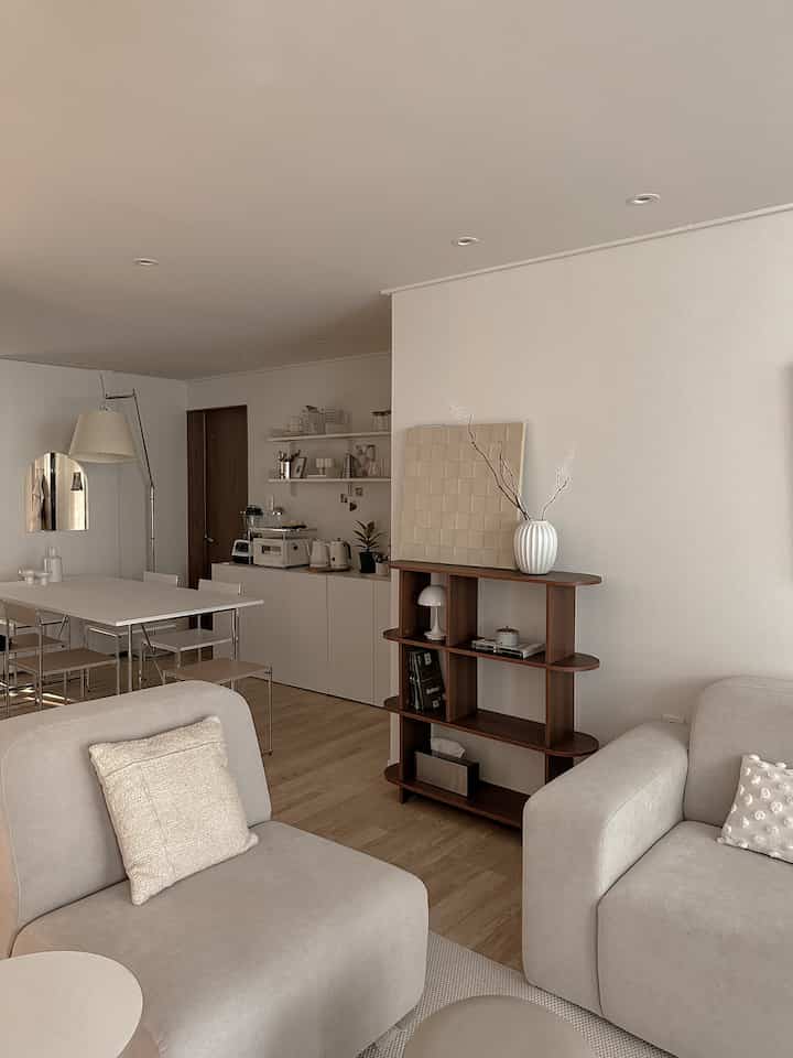 ナチュラルカラーとブラウンのトーンが特徴のリビング、深みのあるウォールナットの棚とベージュのソファが配置されたシンプルで居心地の良い空間