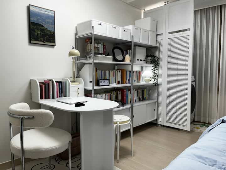White-toned studio study space featuring a modern desk and neatly organized bookshelf creating a book cafe atmosphere